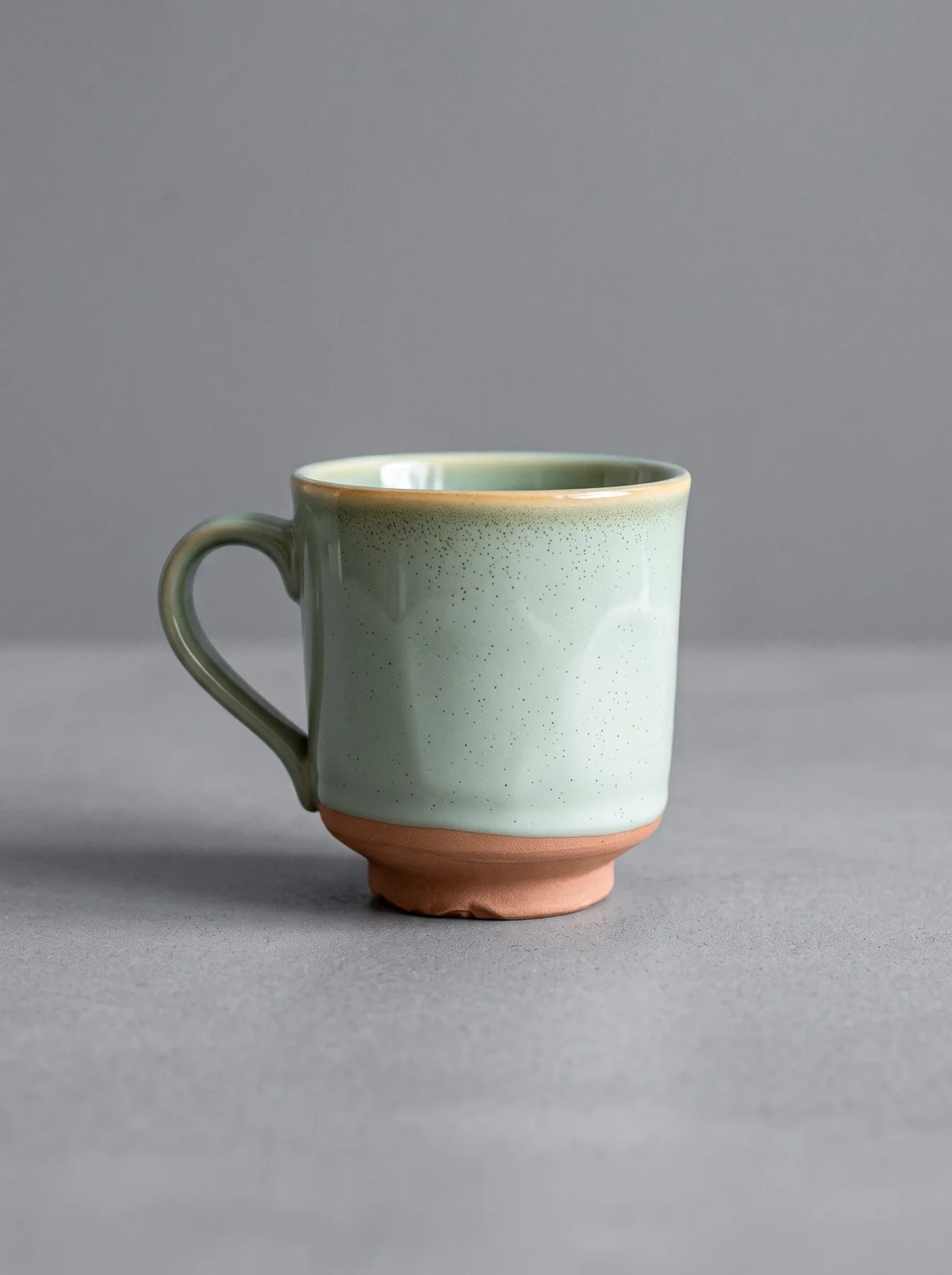 Studio shot: the same sage mug with a terracotta foot on a plain grey background with a soft shadow and no text